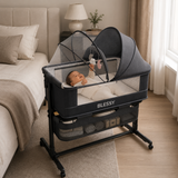 A baby lying on their back inside a BLESSY portable bassinet with a canopy and mesh sides, set in a softly lit bedroom next to a bed.