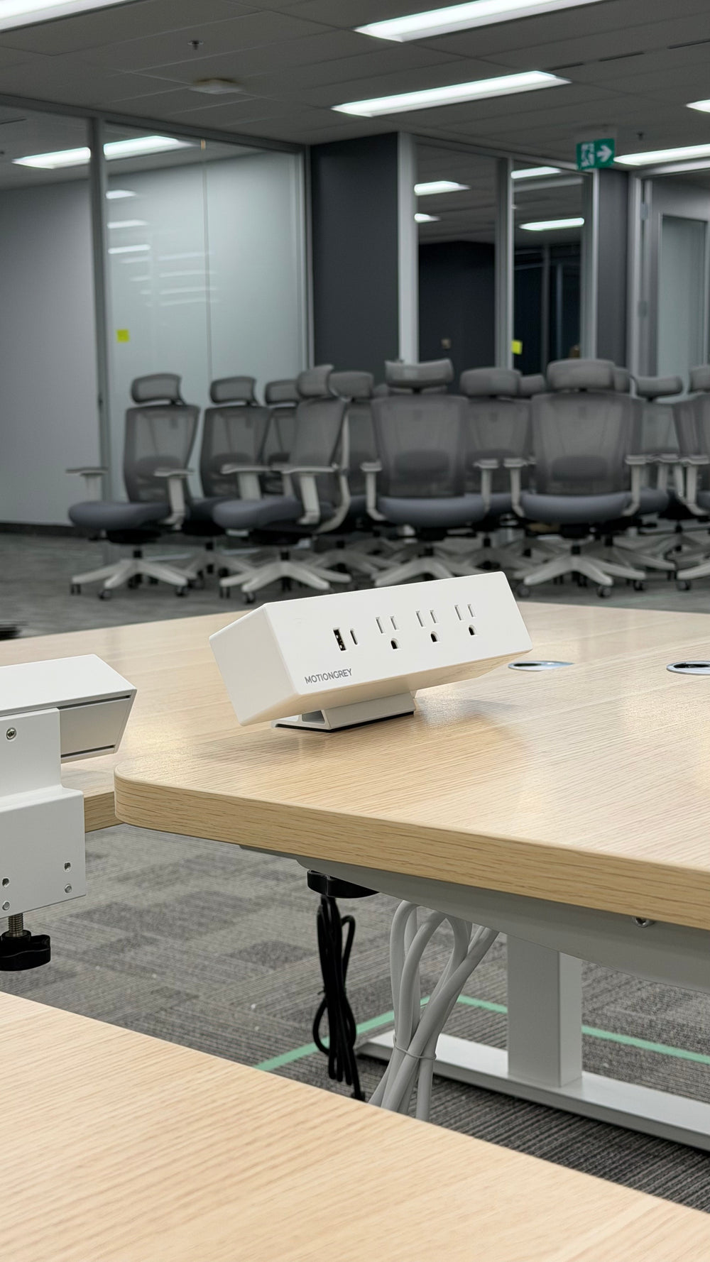 White MotionGrey clamp-on surge protector on a Motion Series desk, with CloudMesh chairs in the background.